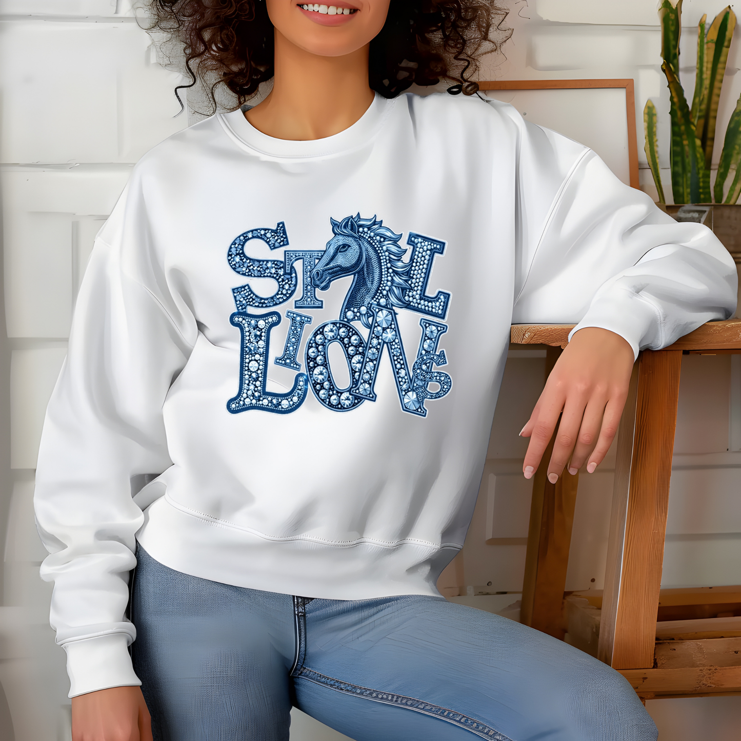 A woman wearing a white crewneck sweatshirt with a blue graphic that reads 'STILL LOVE' in a decorative patterned font, paired with light blue jeans, seated in a home setting.
