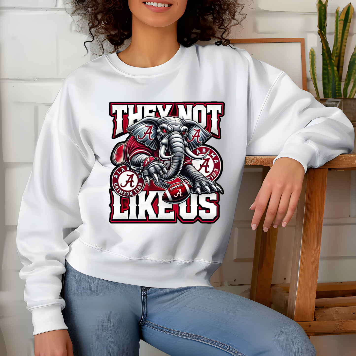 A woman wearing a white crewneck sweatshirt with a graphic of a bulldog and the text 'THEY NOT LIKE US' alongside Alabama Crimson Tide logos, paired with blue jeans.