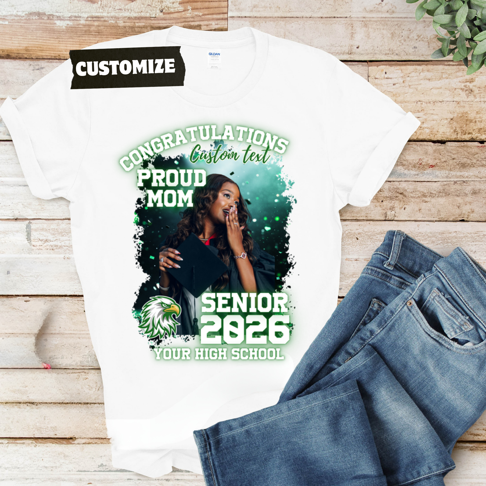 White t-shirt with customizable design for a senior class of 2026, displayed with jeans on a wooden surface.  Customizable graduation photo t-shirt featuring senior portrait, school mascot, and sports organization logos in school colors