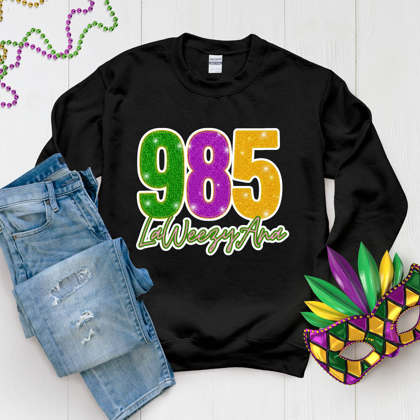 Black sweatshirt with '985 LaWeezyAna' design, paired with denim shorts and a Mardi Gras mask on a light wooden surface.