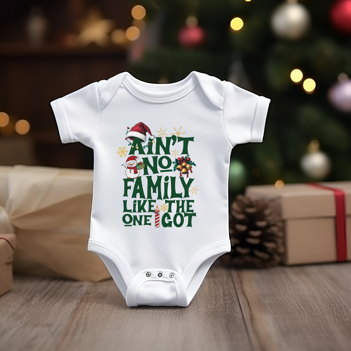 White baby onesie with Christmas-themed text in front of a decorated Christmas tree and presents.