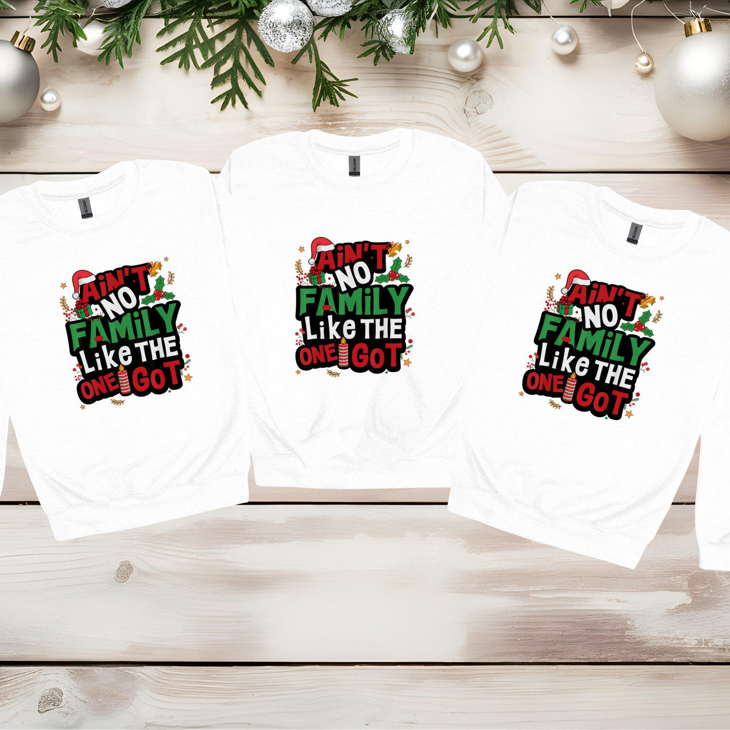 Three white baby onesies with colorful text on a wooden surface with Christmas decorations.
