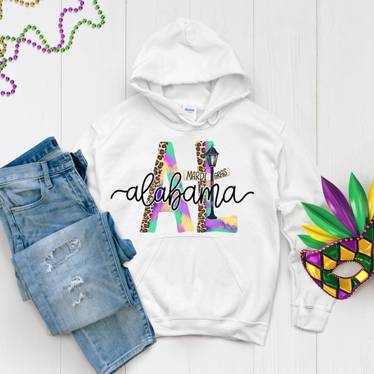 White hoodie with 'Alabama' design, denim shorts, and Mardi Gras mask on a light wooden surface.