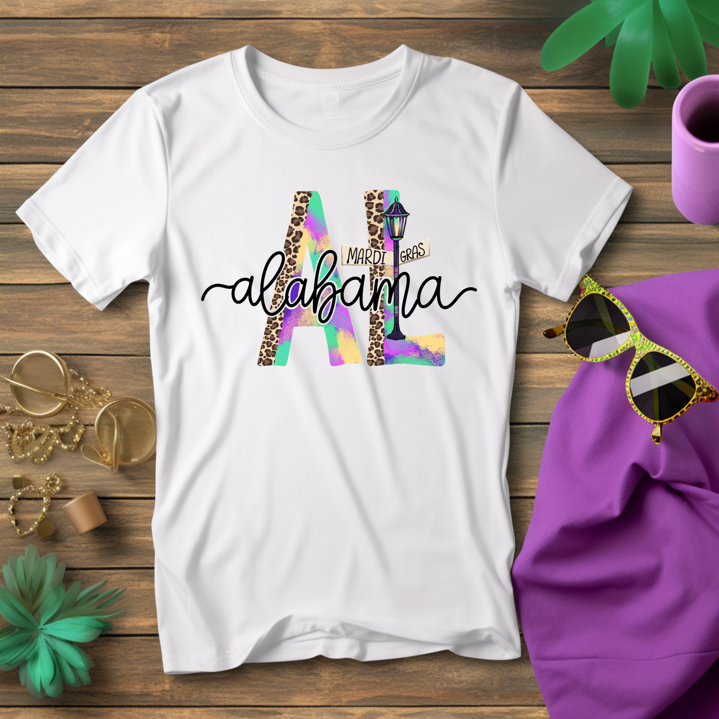 White t-shirt with 'Mardi Gras Alabama' design on a wooden surface with accessories.