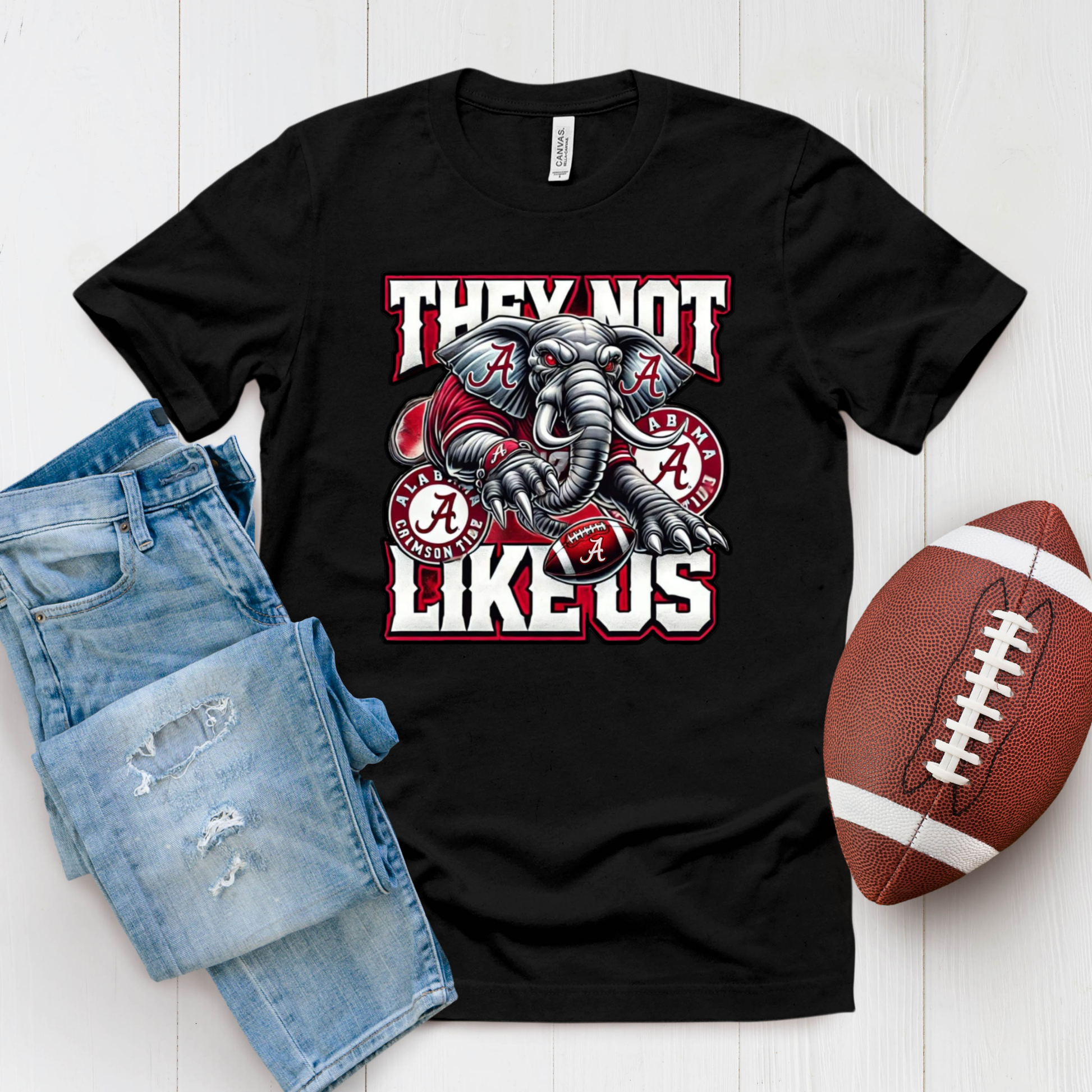 A black graphic t-shirt featuring an Alabama Crimson Tide elephant mascot and the text 'THEY NOT LIKE US' in white and red. The shirt is styled in a flat lay with a pair of distressed blue jeans and a brown football on a white wood background.