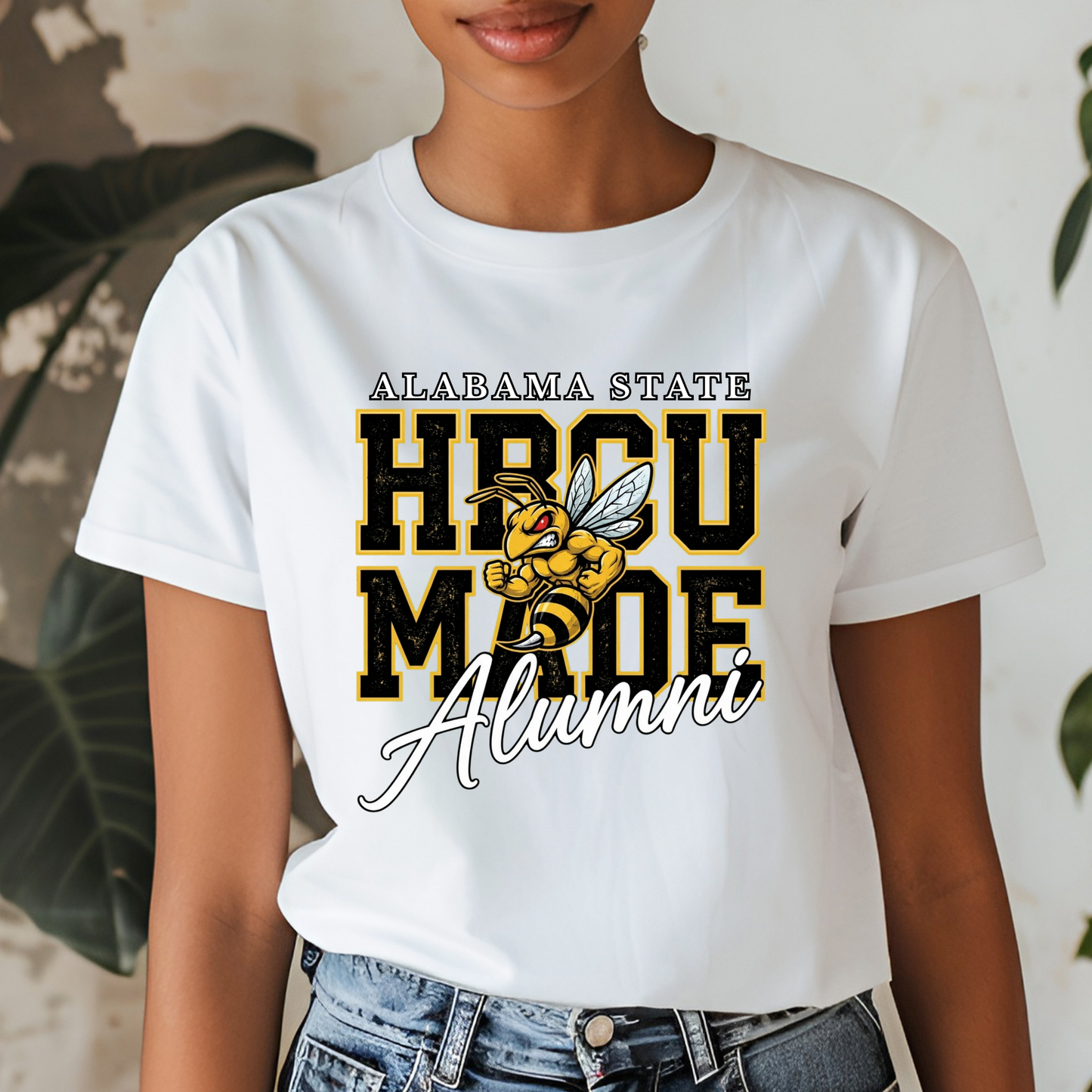 A woman wearing a white graphic t-shirt with 'ALABAMA STATE HBCU MADE Alumni' and a bee mascot printed on the front, paired with blue jeans.