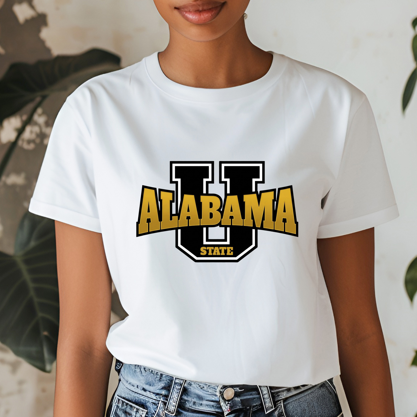 A woman wearing a white short-sleeve graphic T-shirt with a large black and gold 'U Alabama State' logo on the front, paired with blue jeans. The background features a textured light wall and green plant leaves.