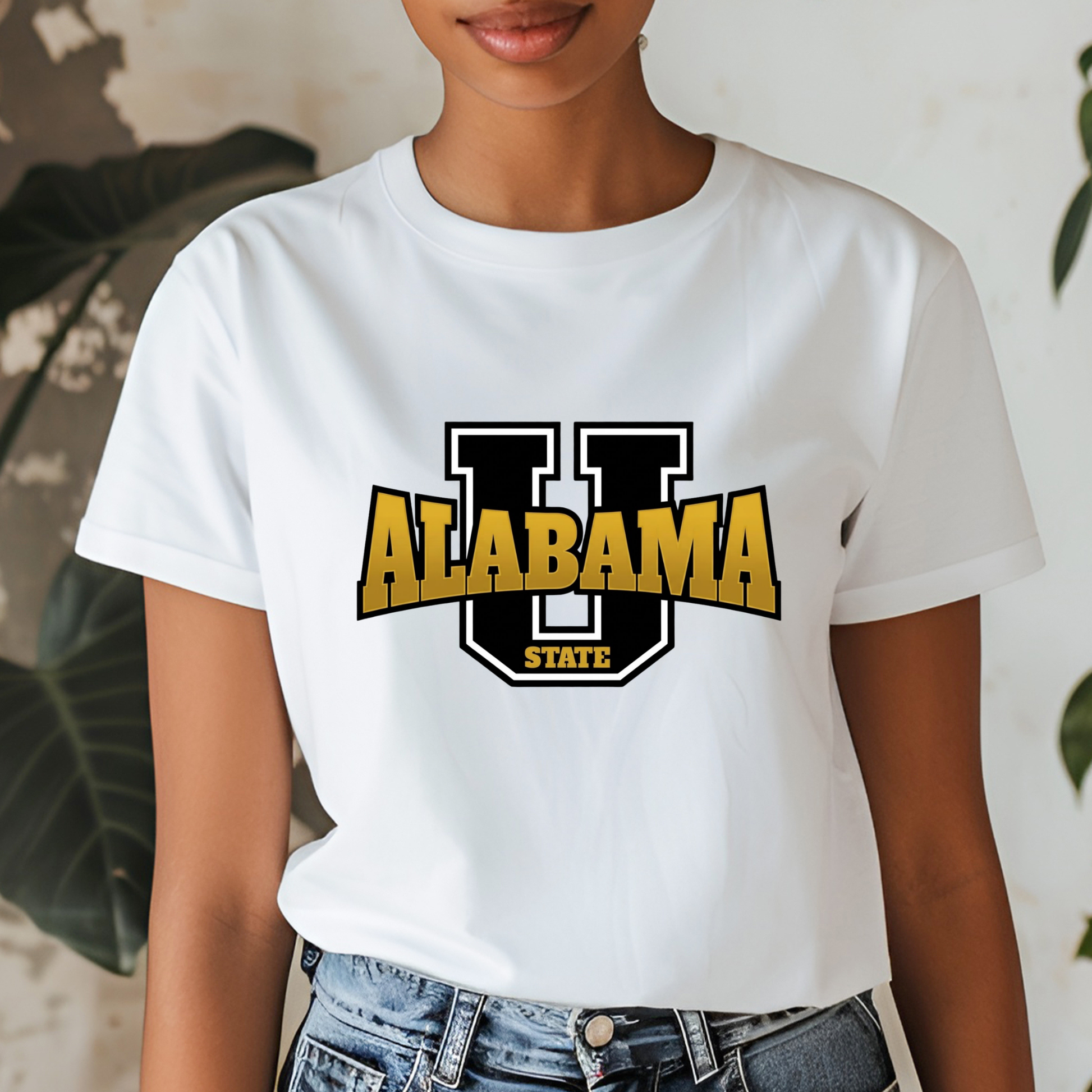 A woman wearing a white short-sleeve graphic T-shirt with a large black and gold 'U Alabama State' logo on the front, paired with blue jeans. The background features a textured light wall and green plant leaves.
