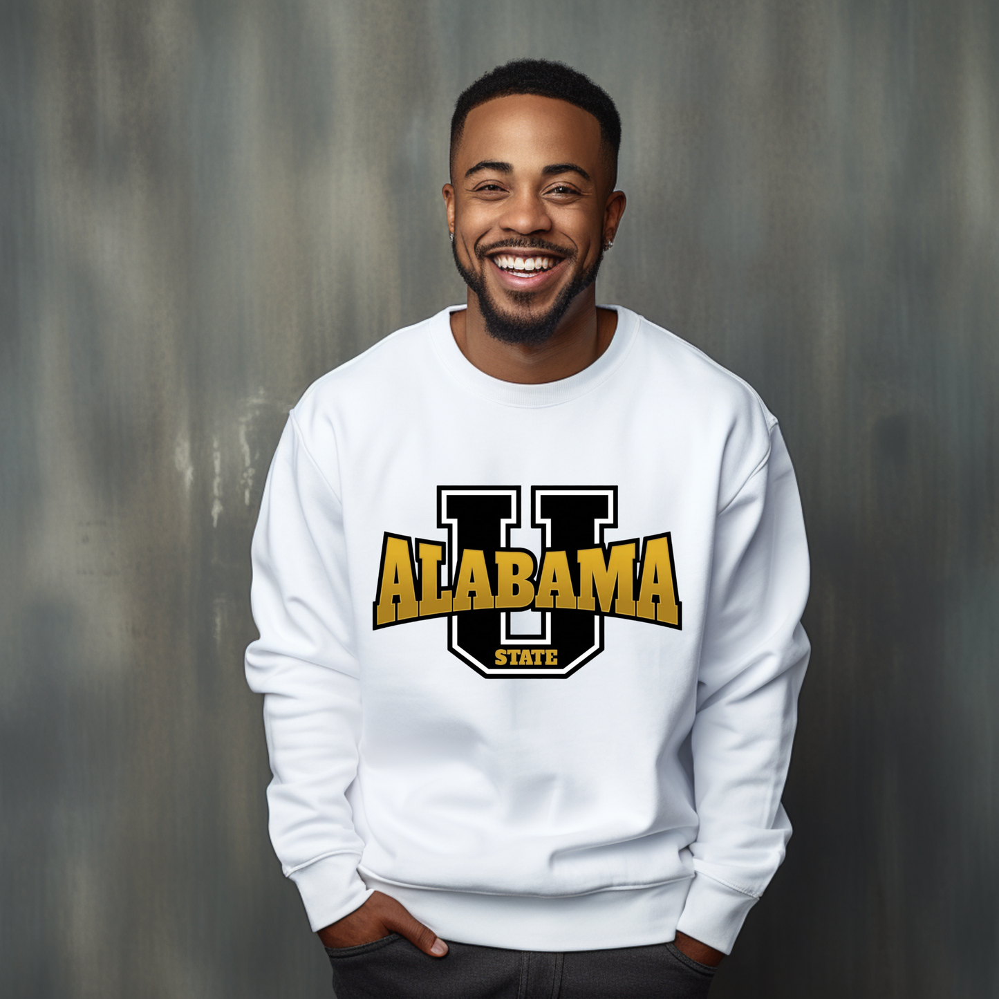 A smiling man wearing a white crewneck sweatshirt with a large 'U ALABAMA STATE' graphic in black and gold on the chest, standing against a textured gray background.
