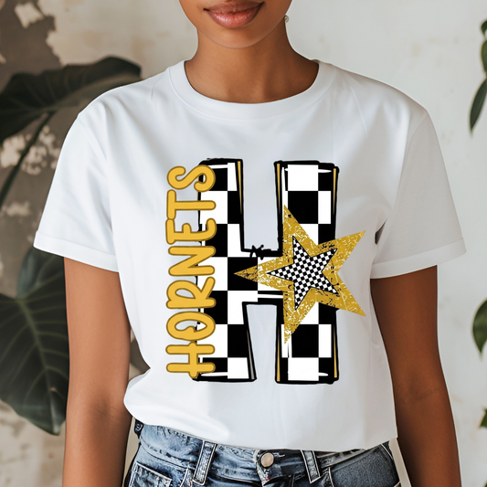 A woman wearing a white graphic t-shirt featuring a large letter 'H' in a black and white checkerboard pattern, a gold glitter star, and the word 'HORNETS' in gold. The photo is cropped from the chin down, showing the model wearing blue jeans against a neutral indoor background with soft leaf shadows.
