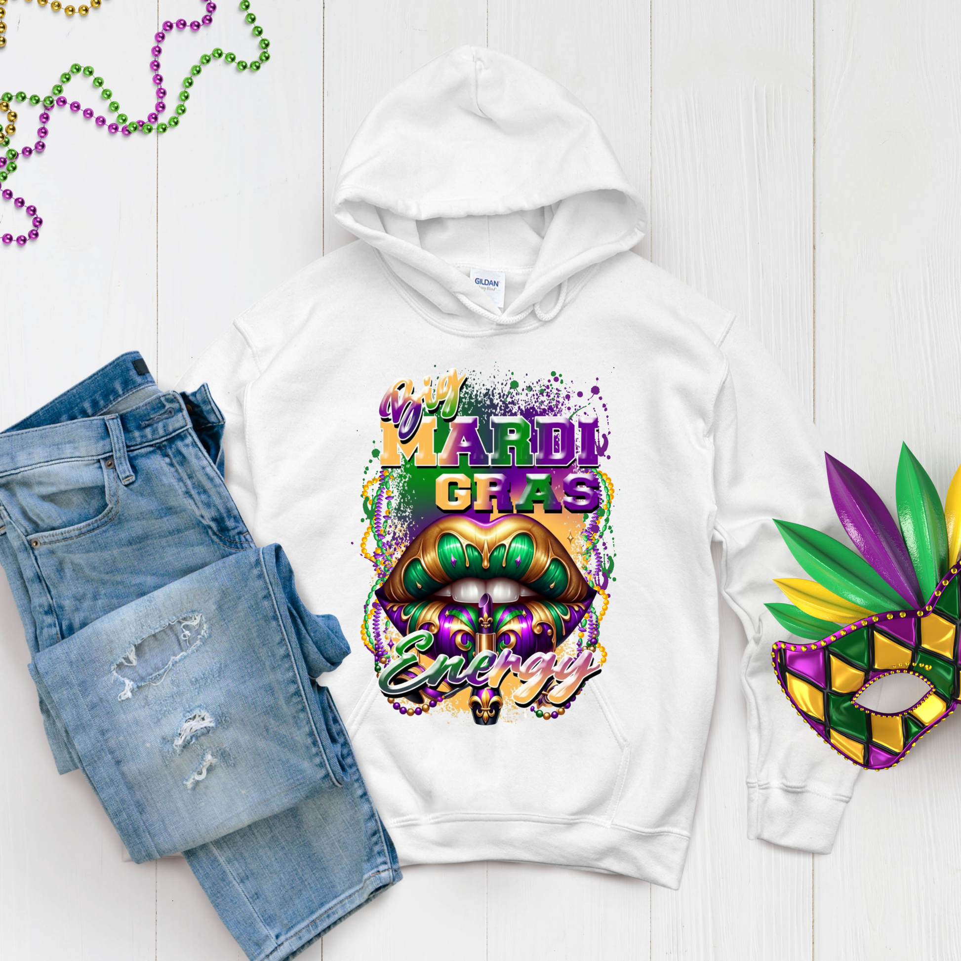 White hoodie with Mardi Gras design, jeans, and mask on a light wooden surface.