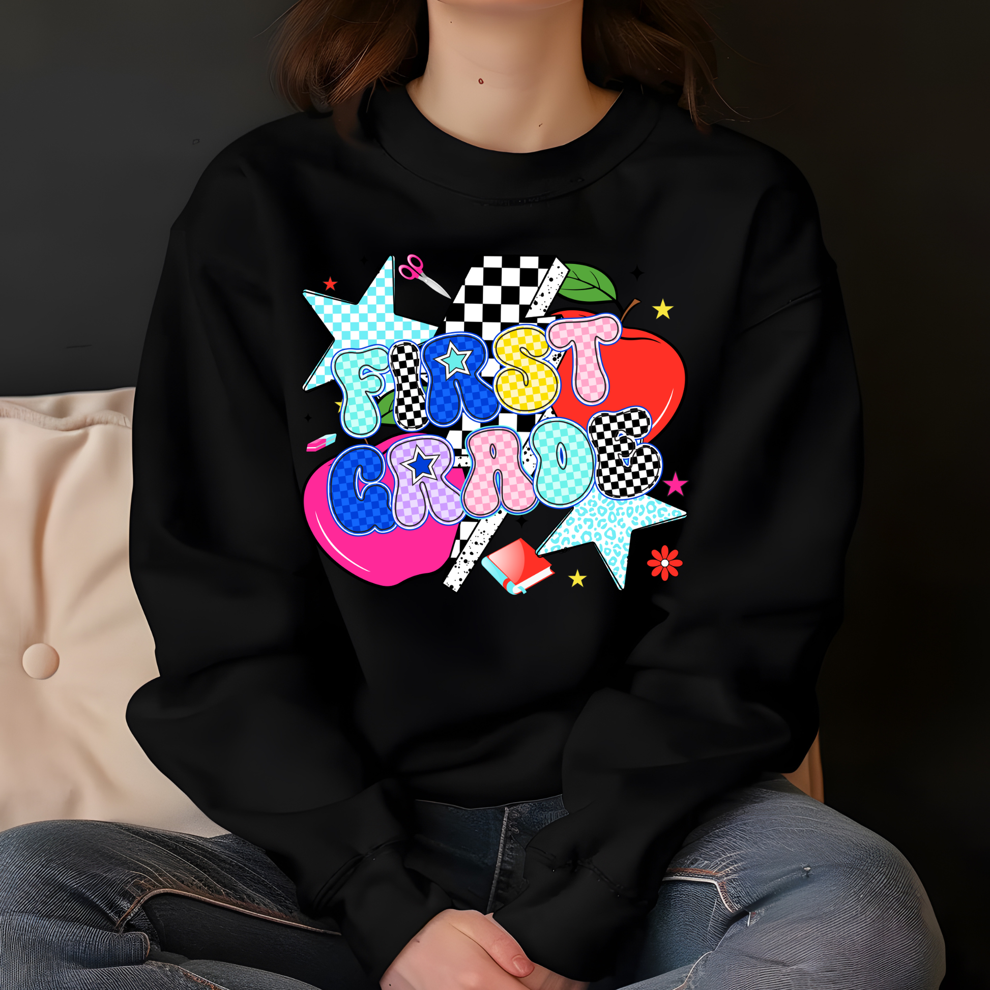 A person wearing a black crewneck sweatshirt with a colorful retro-style graphic that reads 'FIRST CRIO' in pastel bubble letters over a pink heart, star, checkered flag, and other decorative elements. The photo is cropped from the neck down, showing the person seated against a dark background with a beige cushion on the left.
