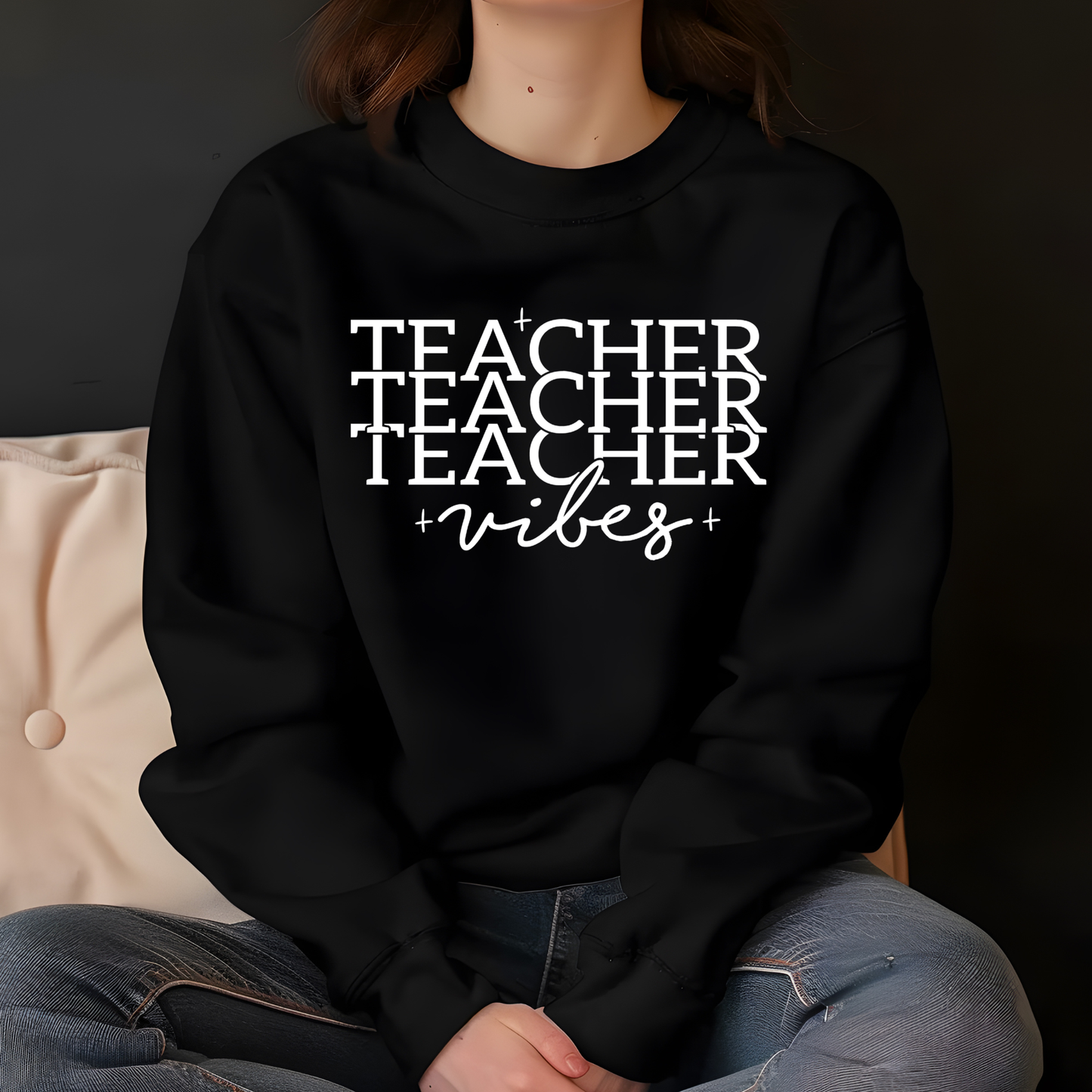 A person wearing a black crewneck sweatshirt with the text 'TEACHER TEACHER TEACHER +vibes+' printed in white across the chest, paired with blue jeans, seated against a dark background.