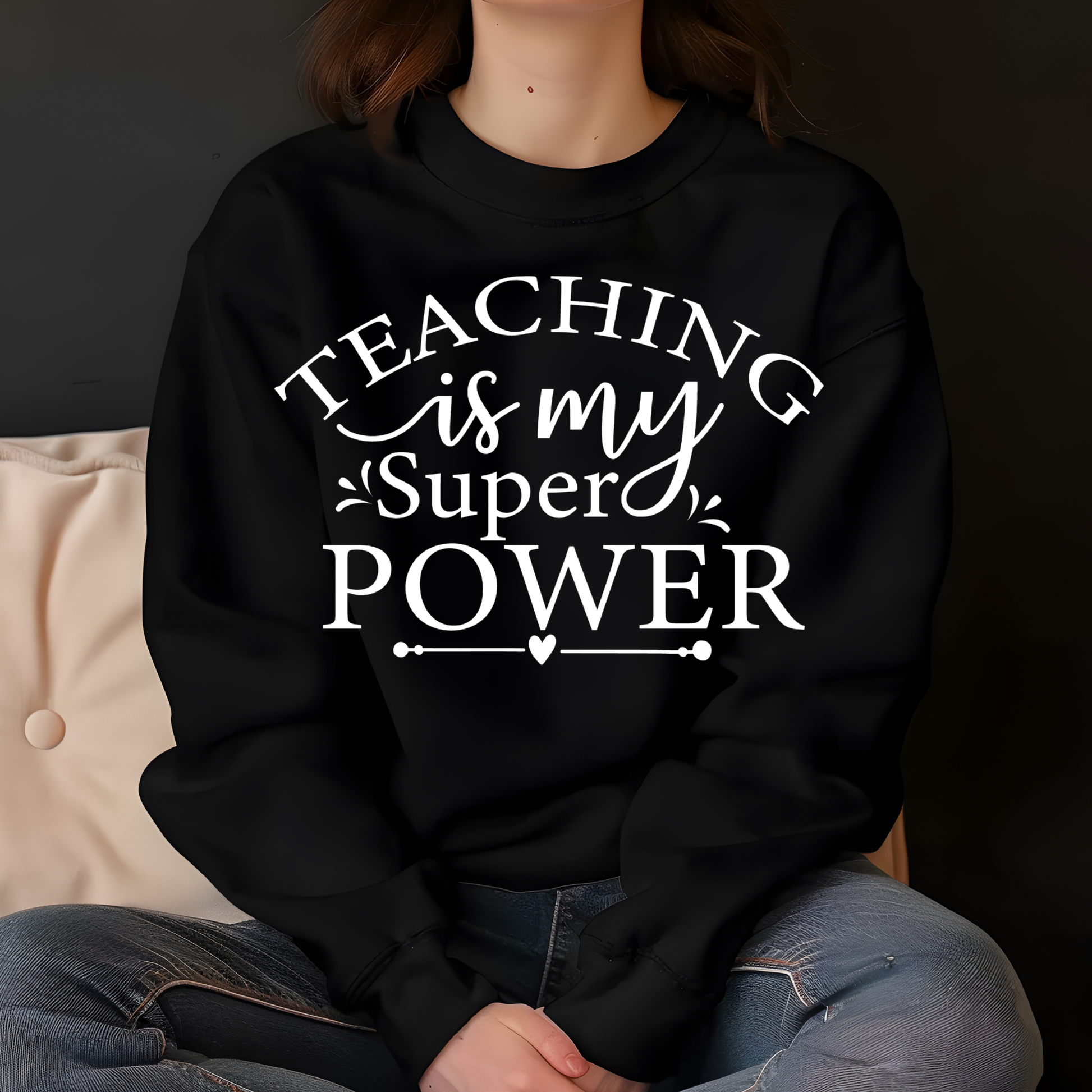 A person wearing a black crewneck sweatshirt with the text 'TEACHING is my Super POWER' printed in white across the chest, paired with blue jeans, seated against a dark background.