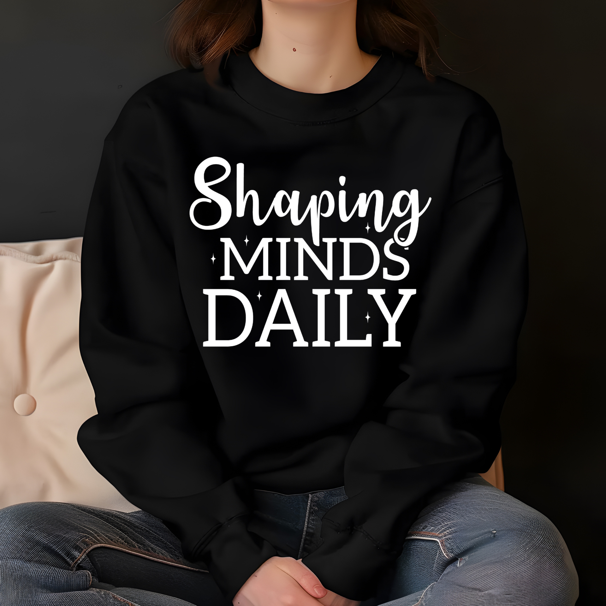 A person wearing a black crewneck sweatshirt with the white text 'Shaping MINDS DAILY' printed on the front, paired with blue jeans, sitting against a dark background.