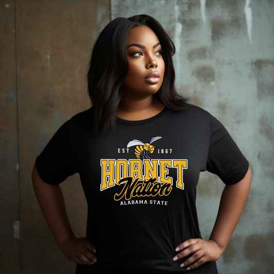 A woman wearing a black short-sleeve graphic T-shirt with a large yellow, white, and gray 'Hornet Nation Alabama State' print on the front, posed against a textured gray wall.