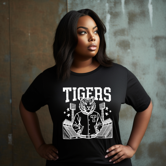A woman wearing a black short-sleeve T-shirt with a white graphic print that reads 'TIGERS' above a tiger mascot in a baseball uniform, posed against a textured gray wall.