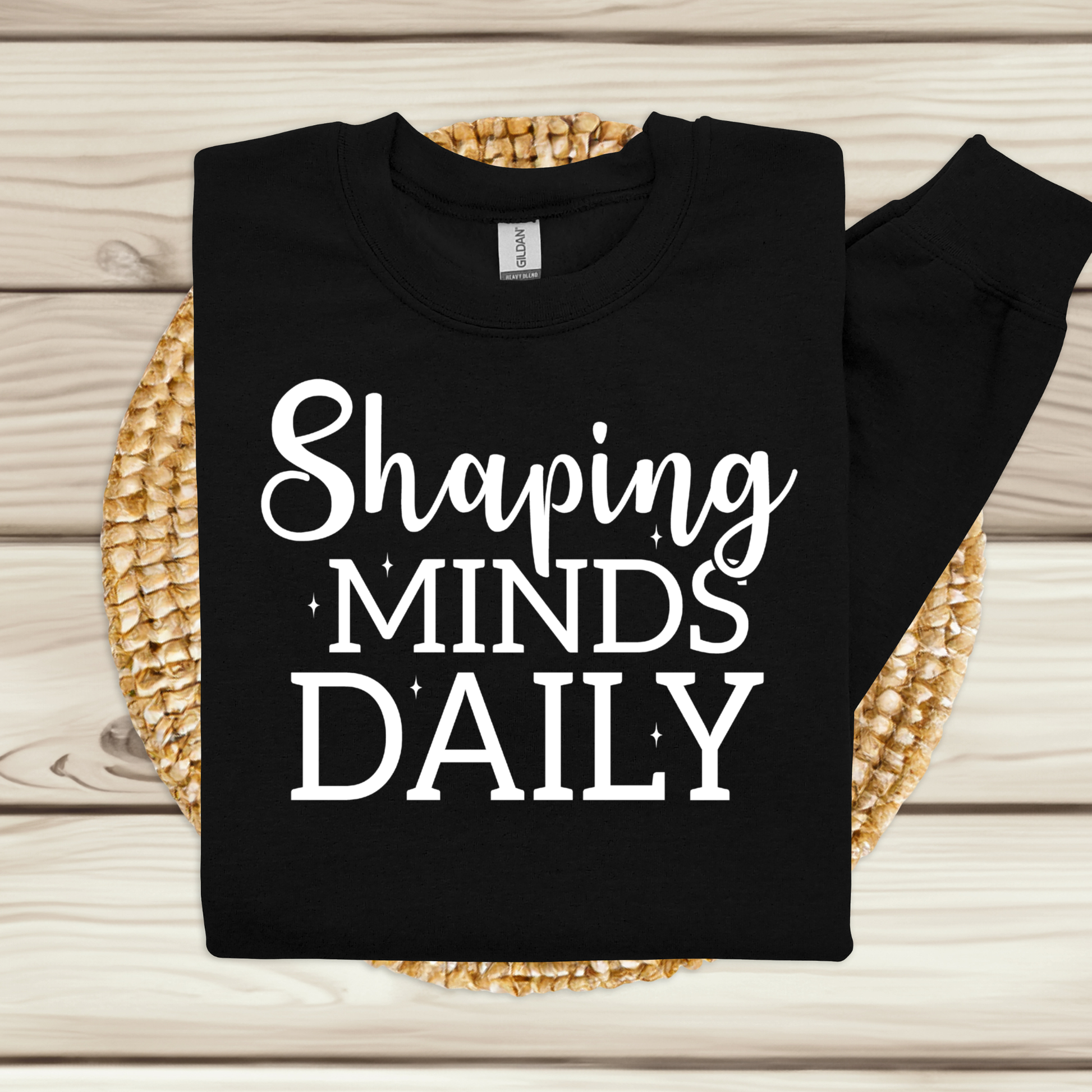 A black long-sleeve sweatshirt with the phrase 'Shaping MINDS DAILY' printed in white across the front, laid flat on a woven straw mat over a light wood plank surface.