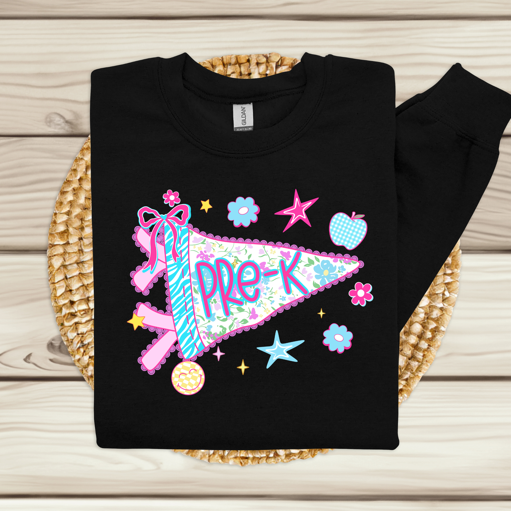 A black long-sleeve sweatshirt laid flat on a woven straw mat over a light wood plank surface. The sweatshirt features a colorful 'Pre-K' pennant graphic with pink, blue, and green floral details, stars, flowers, and a small donut illustration.