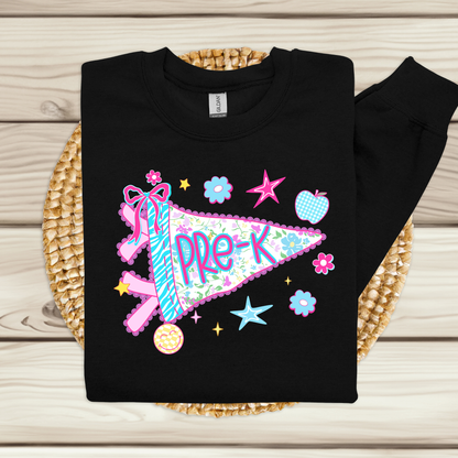 A black long-sleeve sweatshirt laid flat on a woven straw mat over a light wood plank surface. The sweatshirt features a colorful 'Pre-K' pennant graphic with pink, blue, and green floral details, stars, flowers, and a small donut illustration.