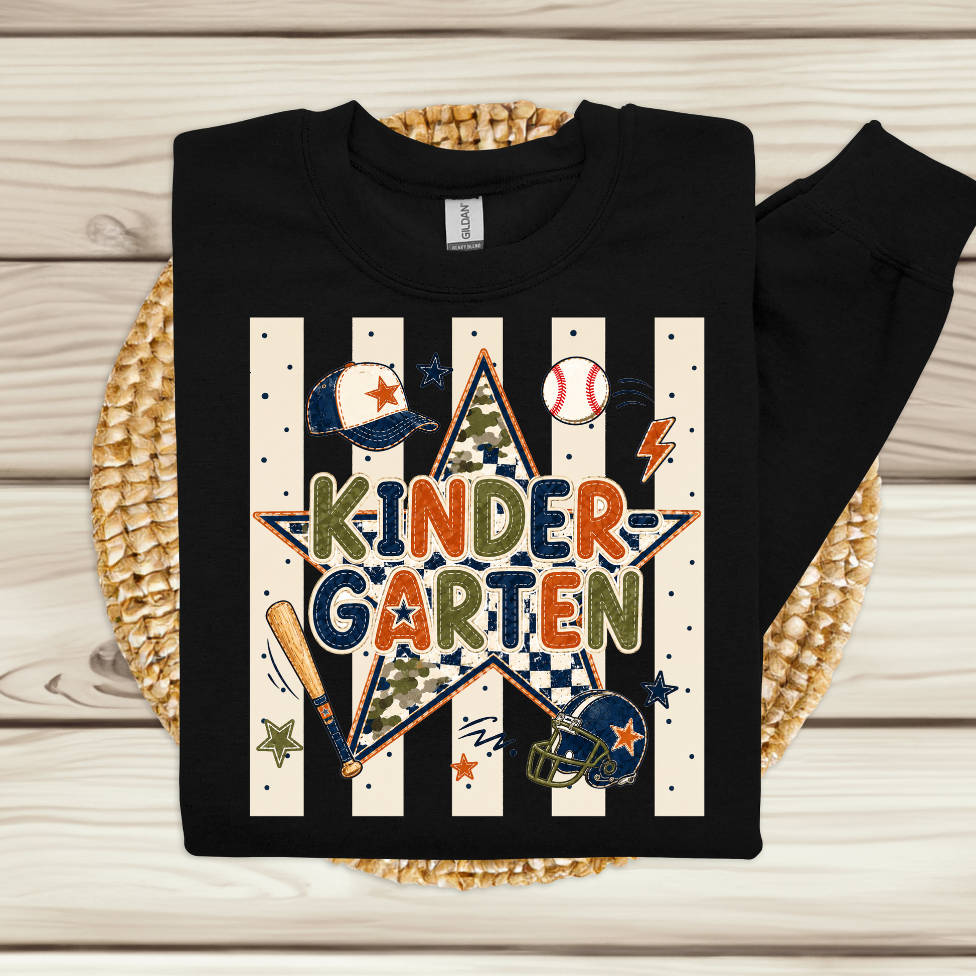 A black long-sleeve sweatshirt laid flat on a woven round mat over a light wood plank background. The sweatshirt features a large graphic design with the words 'KINDER GARTEN' in multicolored letters inside a star, surrounded by baseball and softball themed illustrations including a bat, ball, helmet, and baseball/softball motifs on cream-and-white vertical stripes.