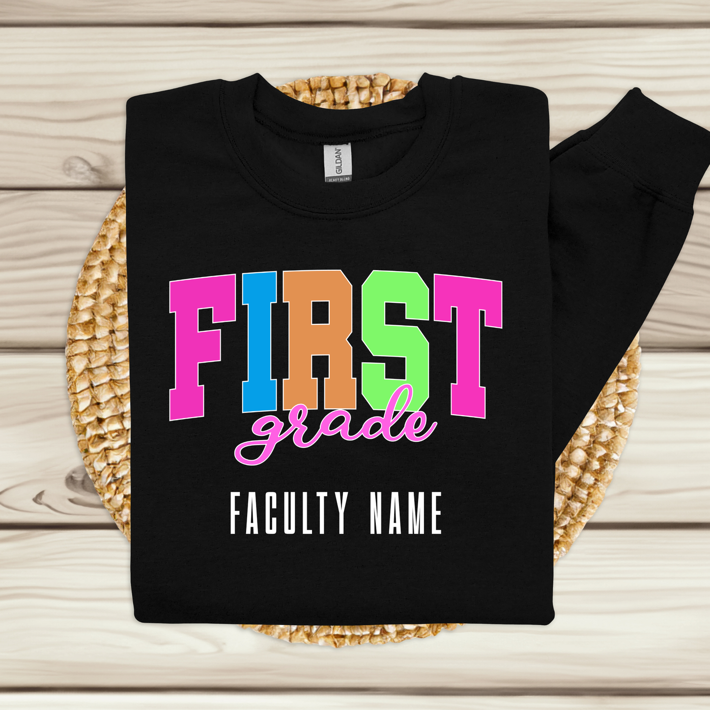 A black long-sleeve sweatshirt laid flat on a woven round mat over a light wood plank surface. The sweatshirt has colorful block letters spelling 'FIRST grade' with 'FACULTY NAME' printed below in white.