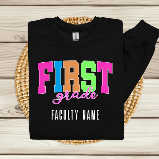 A black long-sleeve sweatshirt laid flat on a woven round mat over a light wood plank surface. The sweatshirt has colorful block letters spelling 'FIRST grade' with 'FACULTY NAME' printed below in white.