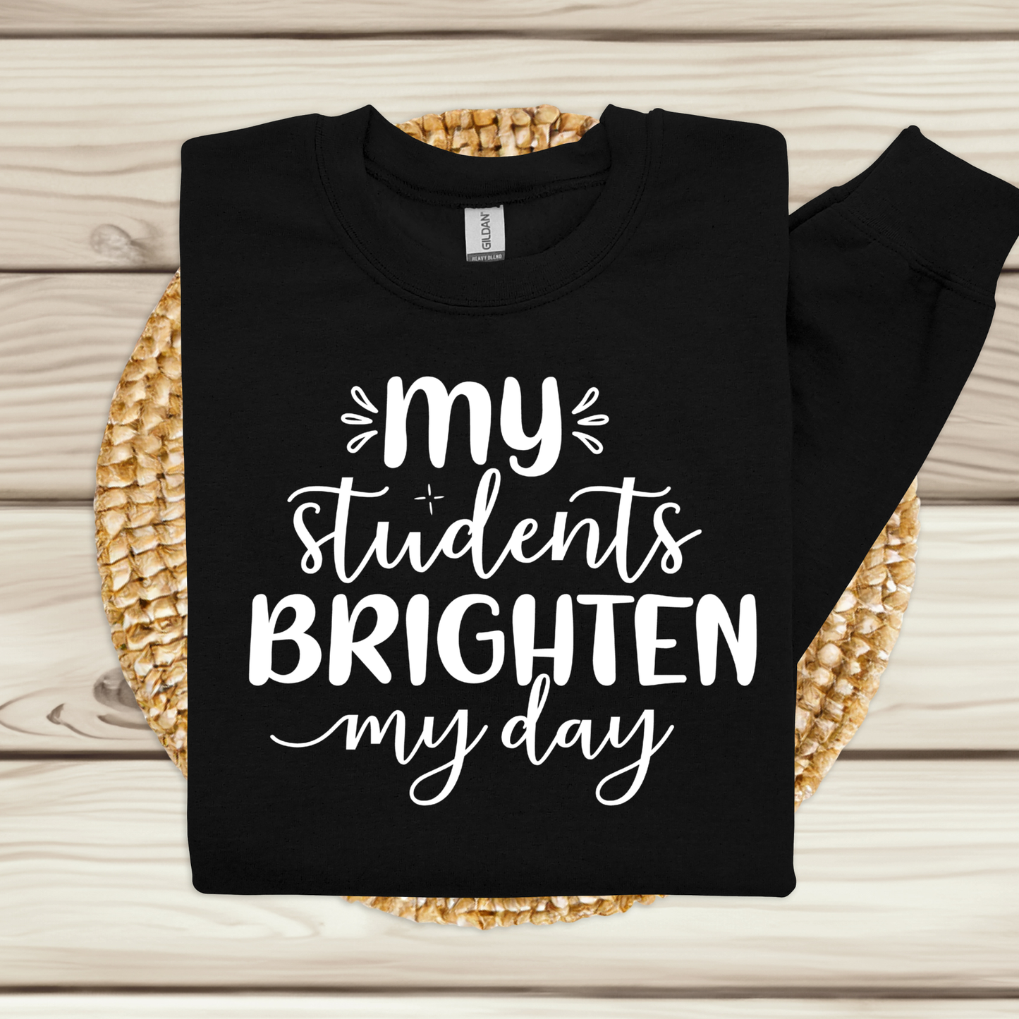 A black long-sleeve crewneck sweatshirt with white text reading 'my students BRIGHTEN my day' is displayed flat on a light wood background, resting on a round woven straw mat.