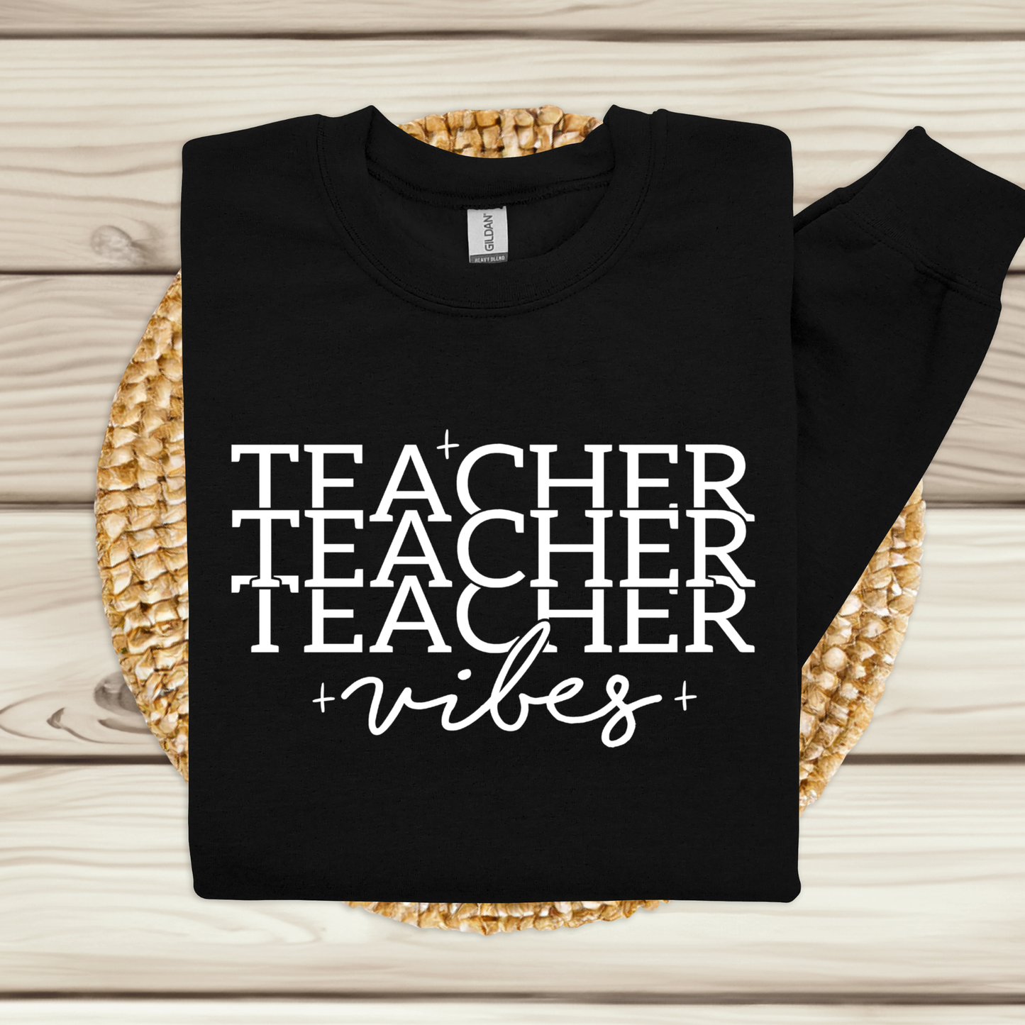 A black long-sleeve crewneck sweatshirt with 'TEACHER TEACHER TEACHER vibes' printed in white text, laid flat on a woven round mat over a light wood plank background.