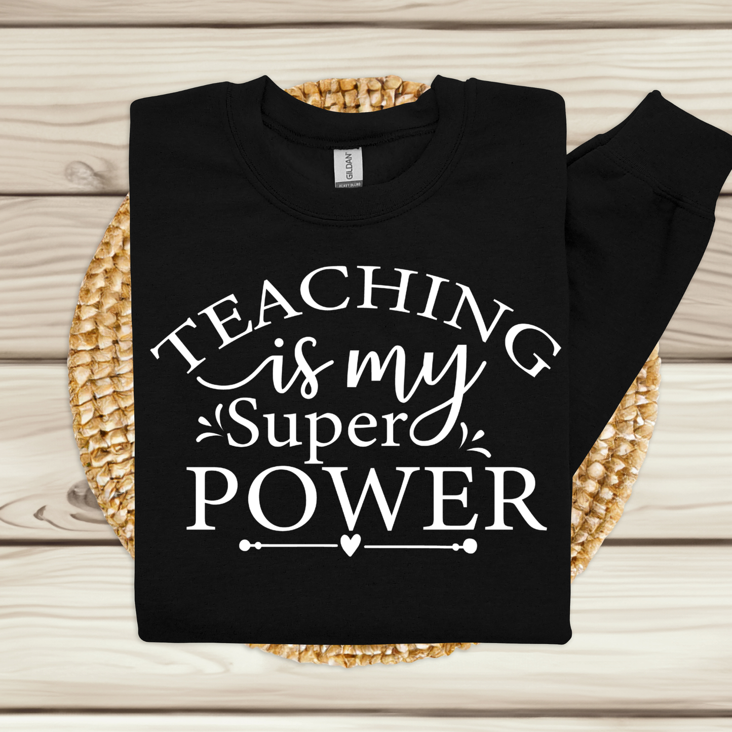 A black long-sleeve t-shirt with white text reading 'TEACHING is my Super POWER' is laid flat on a round woven placemat over a light wood plank background.