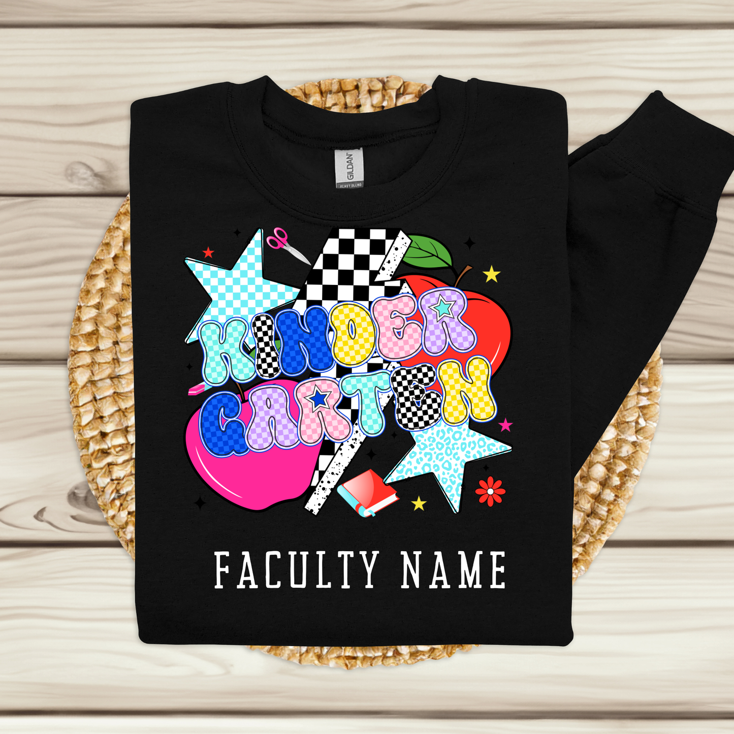 A black long-sleeve sweatshirt laid flat on a woven round mat over a light wood plank surface. The front has a colorful playful graphic reading 'KINDER CARTON' with stars, hearts, checkered details, and a red apple, plus white text reading 'FACULTY NAME' below.