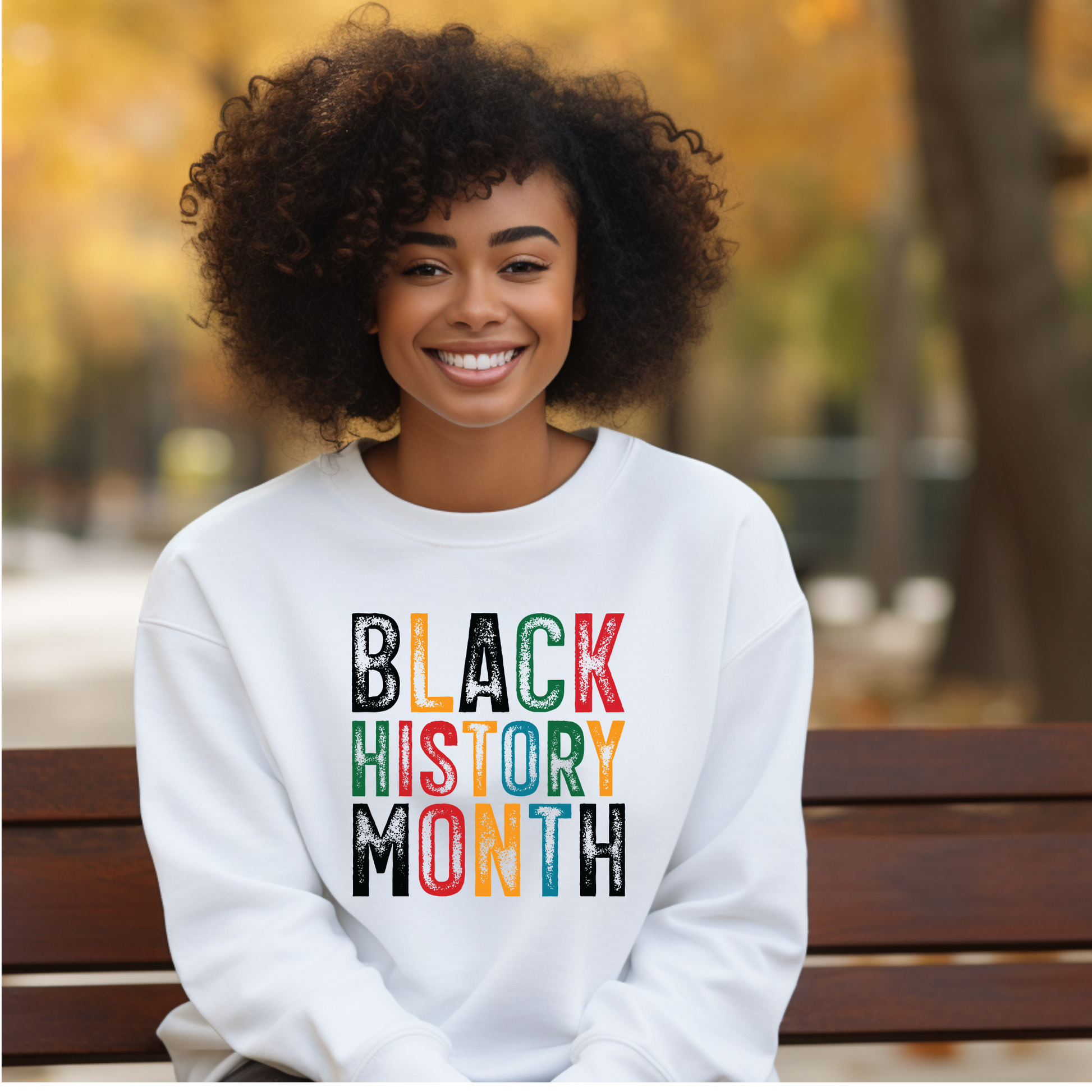 Person wearing a white sweatshirt with 'BLACK HISTORY MONTH' in colorful letters, sitting on a bench outdoors.