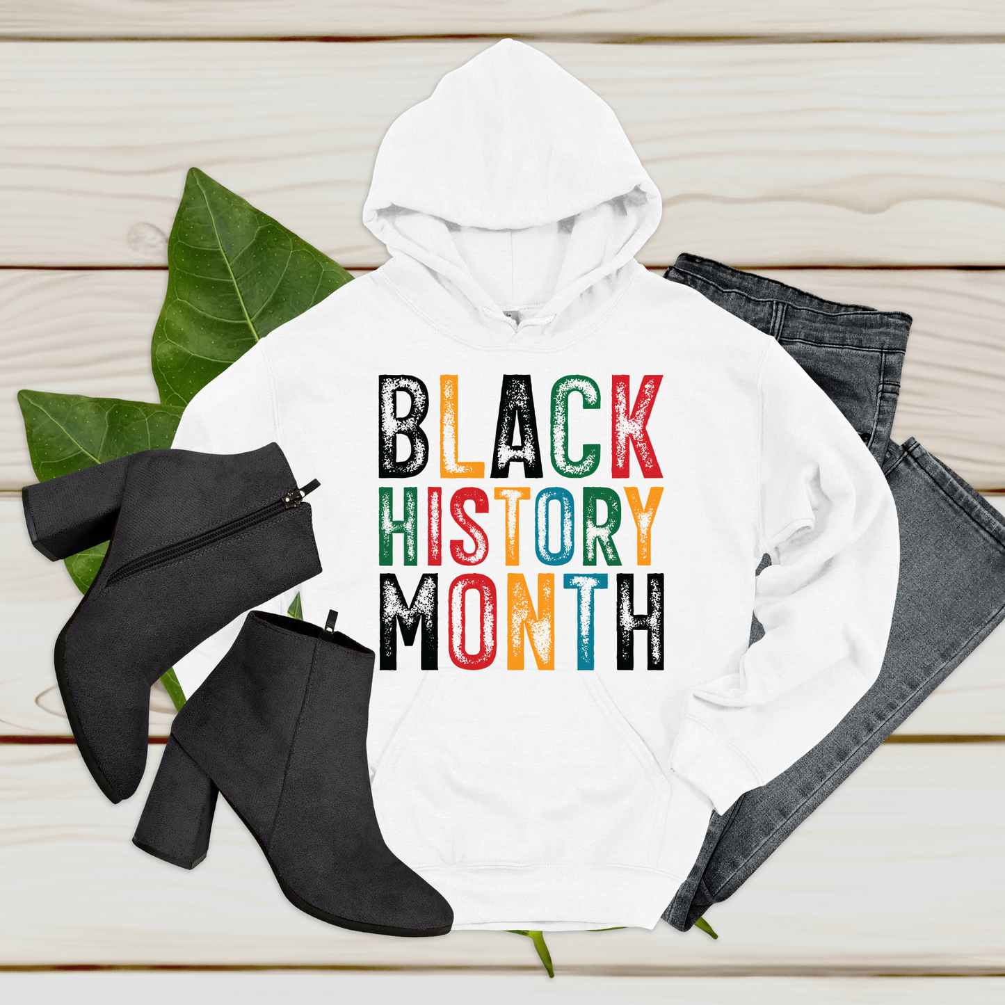 White hoodie with 'Black History Month' text, black ankle boots, and gray jeans on a wooden surface.