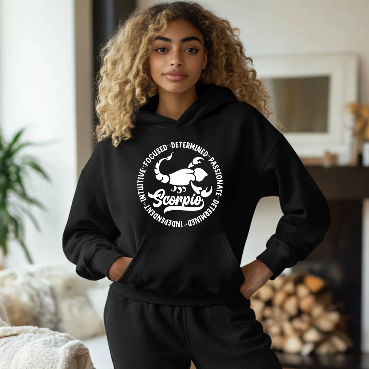 Woman wearing a black hoodie featuring a white circular Scorpio zodiac graphic with a scorpion illustration, the word "Scorpio," and trait keywords like intuitive, focused, determined, passionate, and independent