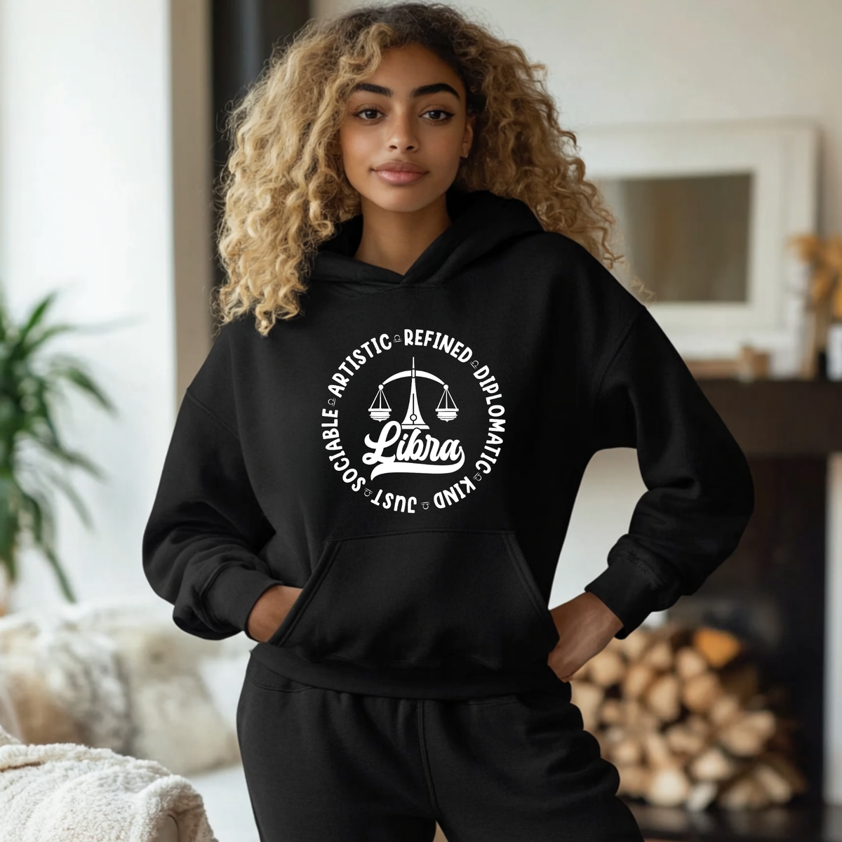 A woman with curly blonde hair wearing a black hoodie and matching sweatpants, standing in a cozy indoor setting with a fireplace and plant in the background. The hoodie features a white graphic of a scale of justice with the word 'Libra' in the center and text around it.