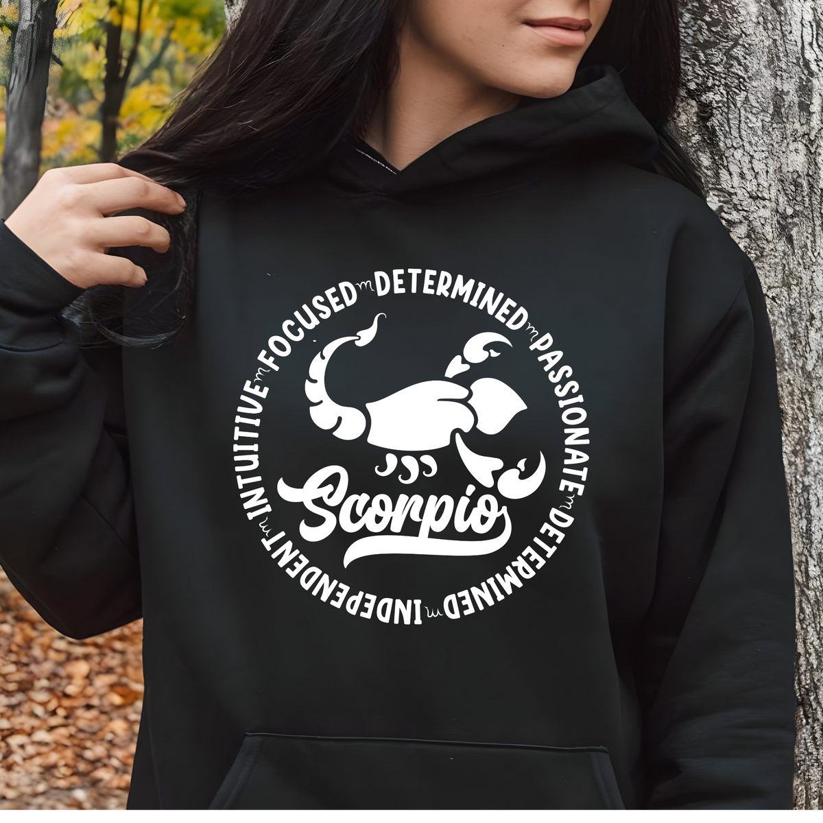 Woman wearing a black crewneck sweatshirt featuring a white circular Scorpio zodiac graphic with a scorpion illustration, the word "Scorpio," and trait keywords like intuitive, focused, determined, passionate, and independent.