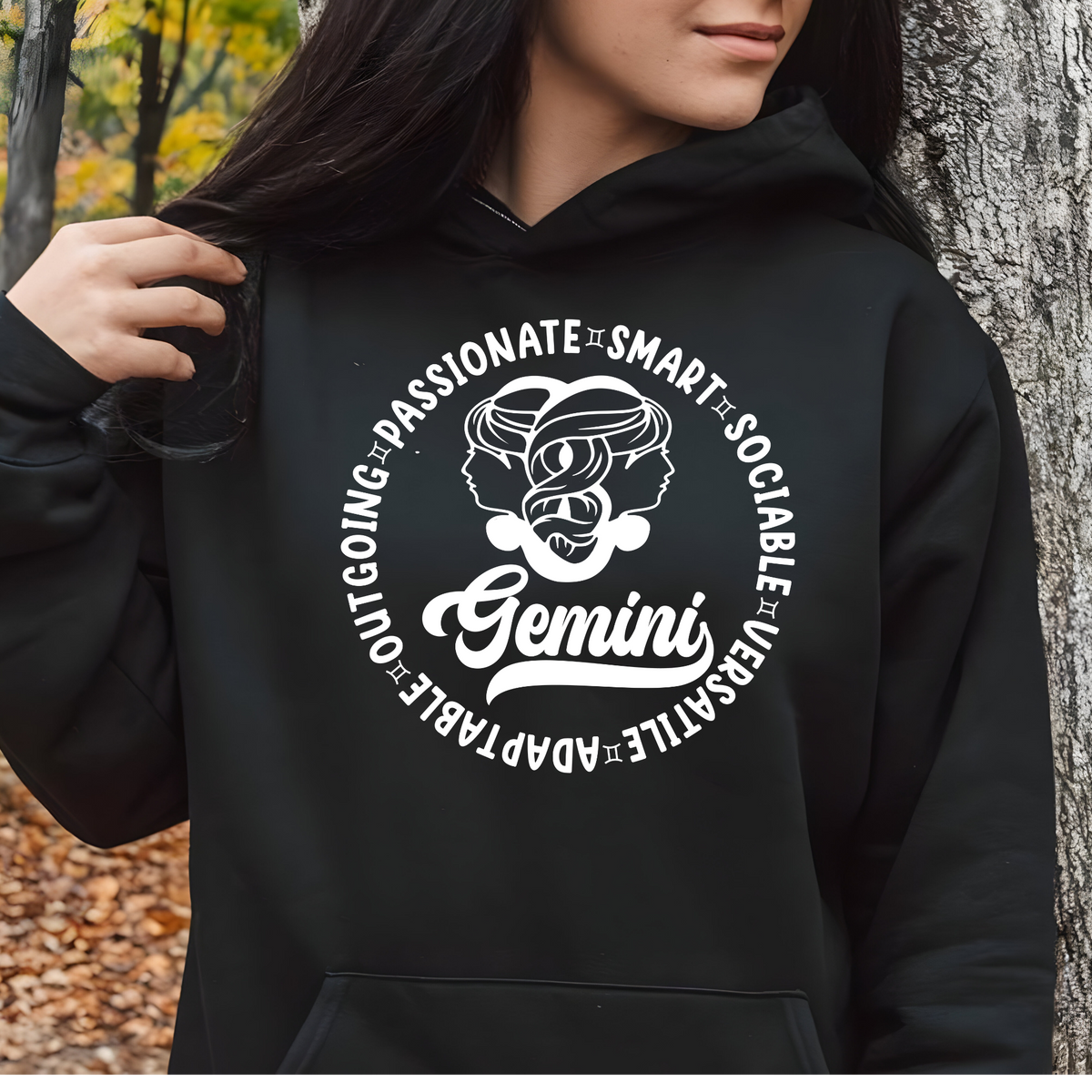 A person wearing a black pullover hoodie with a large white Gemini zodiac graphic on the chest, standing outdoors next to a tree trunk.