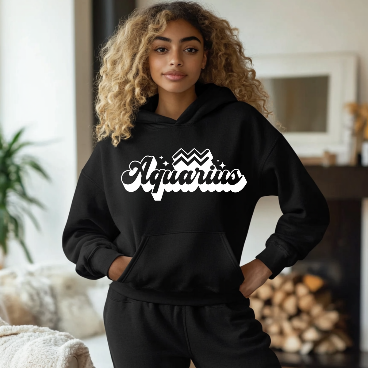 A woman with curly blonde hair wearing a black hoodie and matching sweatpants, featuring a white 'Aquarius' graphic on the chest, posed in a cozy indoor living room setting.