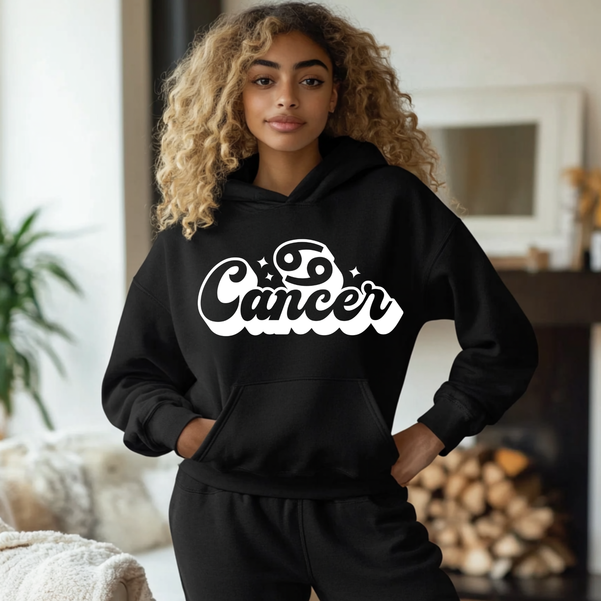 A woman with curly blonde hair wearing a black hoodie with a white 'Cancer' zodiac graphic on the chest, standing in a cozy indoor room with a fireplace in the background.