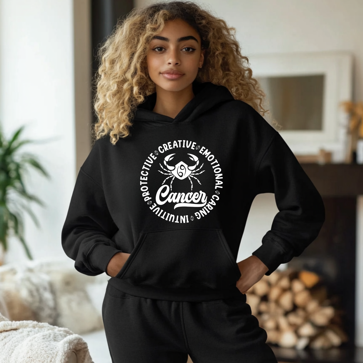A woman with curly blonde hair wearing a black hoodie and matching sweatpants, standing in a cozy indoor setting with a fireplace and plant in the background. The hoodie features a white circular graphic with a scorpion illustration and the word 'Scorpio' surrounded by adjectives.