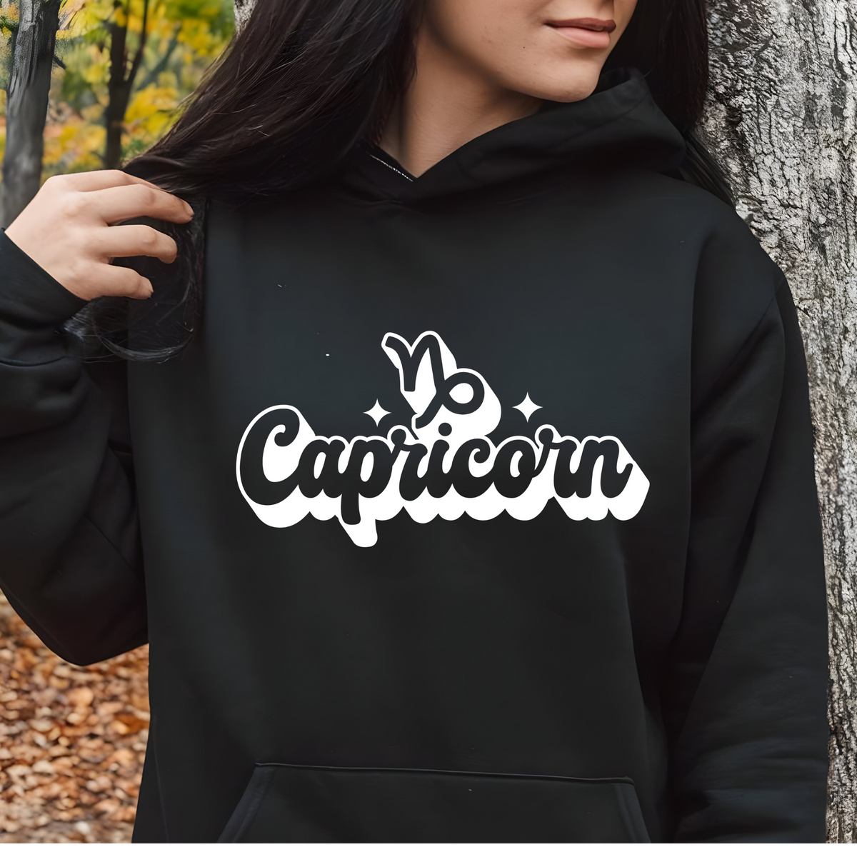 A person wearing a black hoodie with a white 'Capricorn' graphic on the chest, standing outdoors next to a tree trunk.
