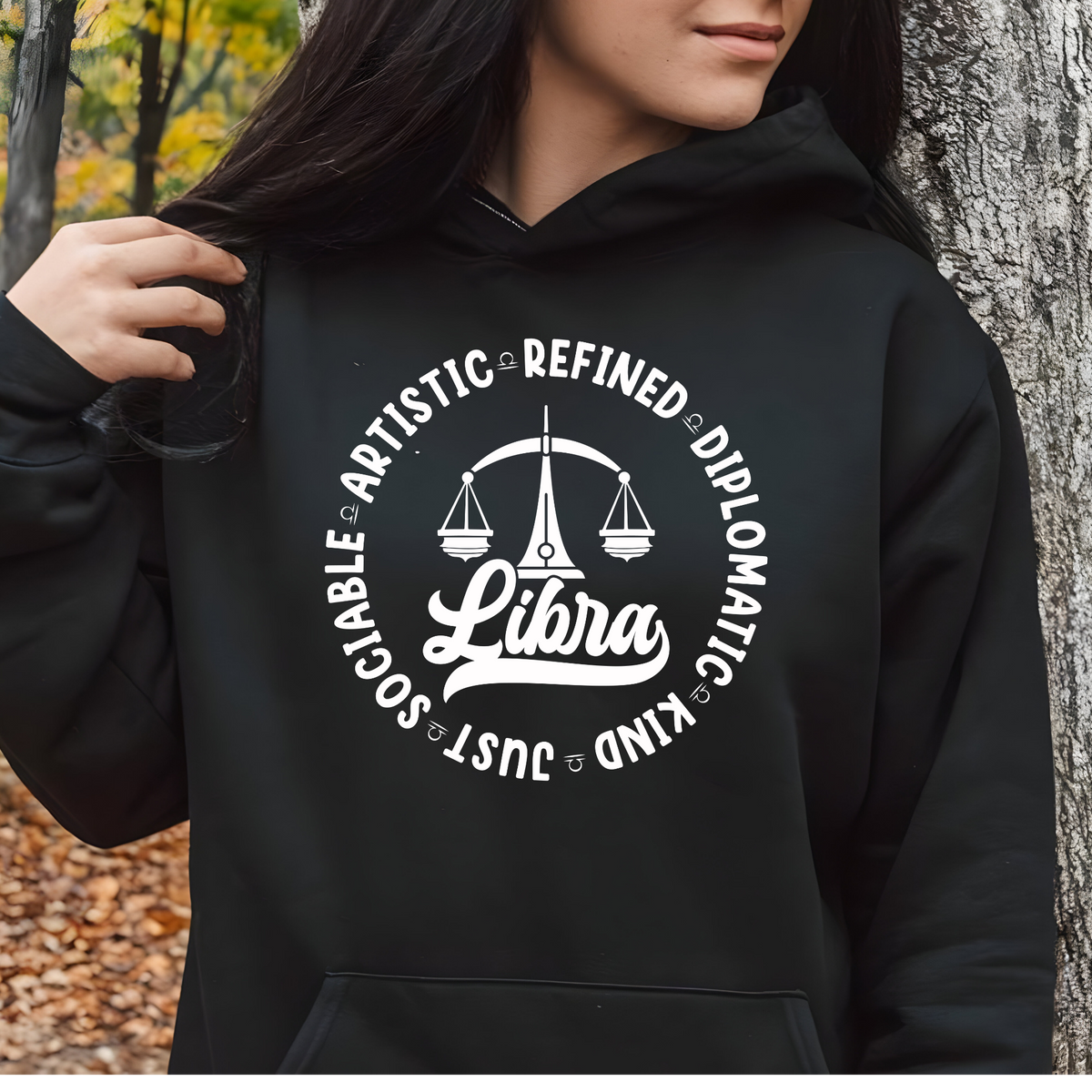 Woman wearing a white crewneck t-shirt featuring a black circular Libra zodiac graphic with a scales illustration, the word "Libra," and trait keywords like artistic, refined, diplomatic, kind, just, and sociable.