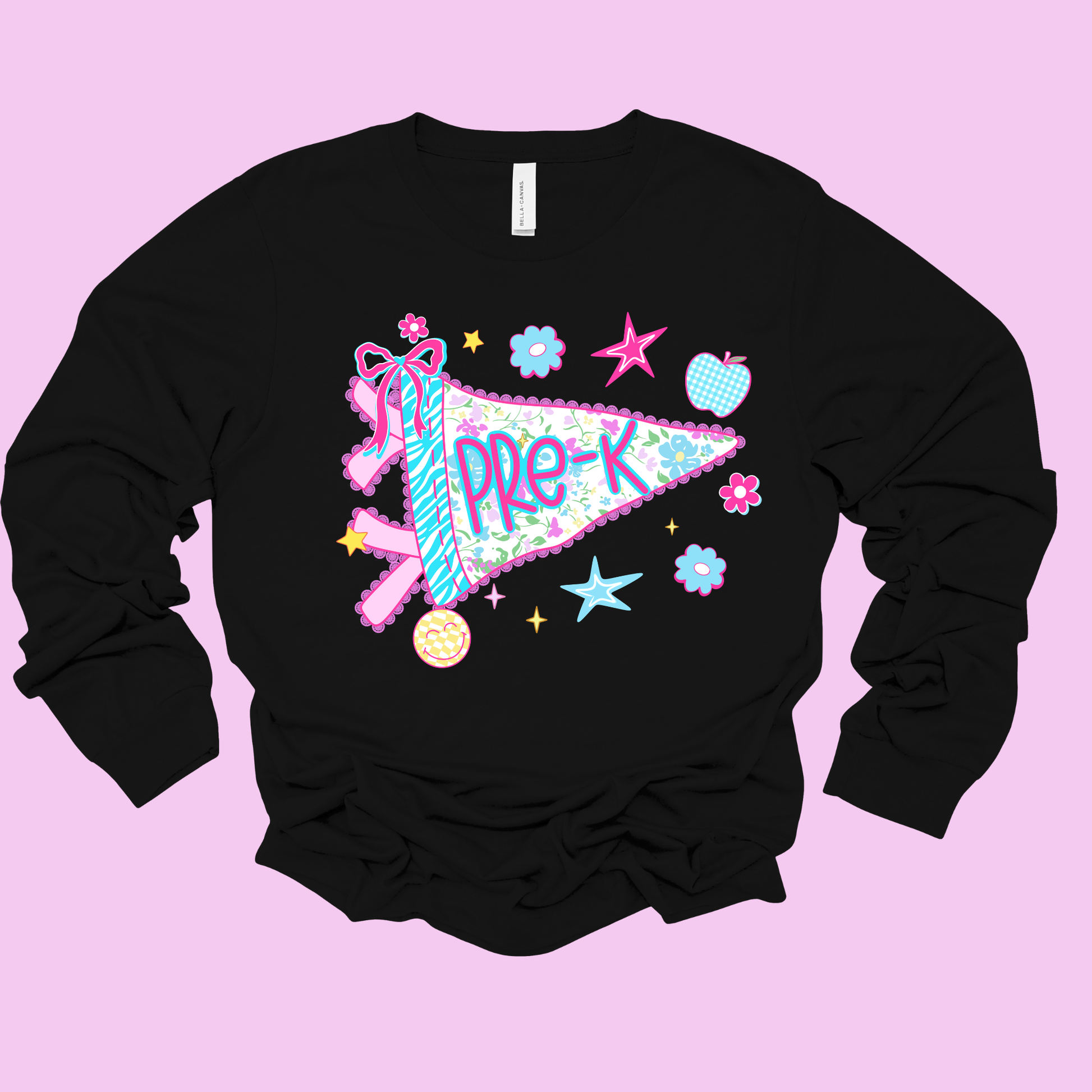 A black long-sleeve crewneck sweatshirt featuring a colorful graphic of a pink and blue patterned pennant with the text 'PRE-K' in purple, surrounded by pastel stars, flowers, and a pink ribbon, displayed on a solid light pink background.