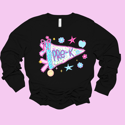 A black long-sleeve crewneck sweatshirt featuring a colorful graphic of a pink and blue patterned pennant with the text 'PRE-K' in purple, surrounded by pastel stars, flowers, and a pink ribbon, displayed on a solid light pink background.
