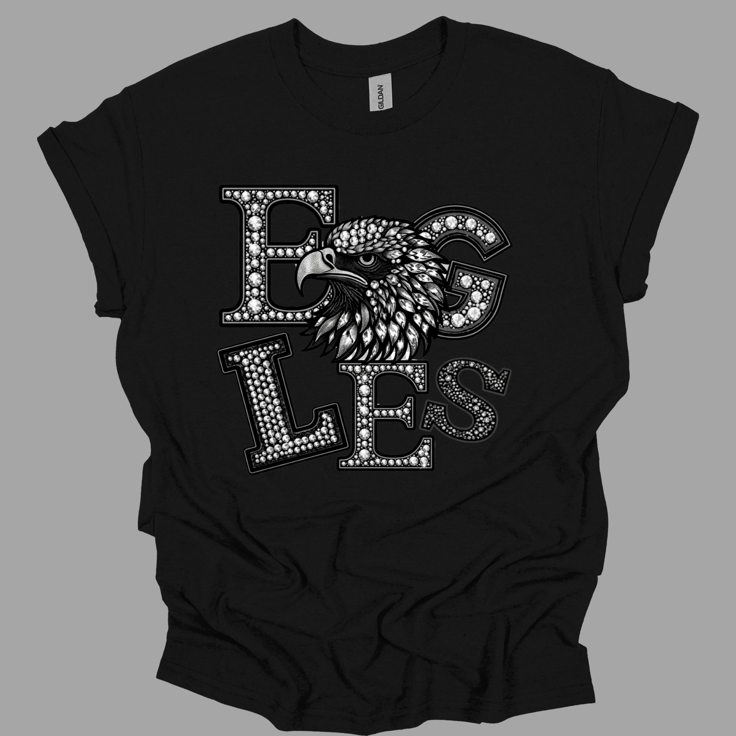 A black short-sleeve t-shirt with a graphic print of the word 'EGGS' in a rhinestone-style font, featuring an eagle's head integrated into the design, shown on a solid gray background.