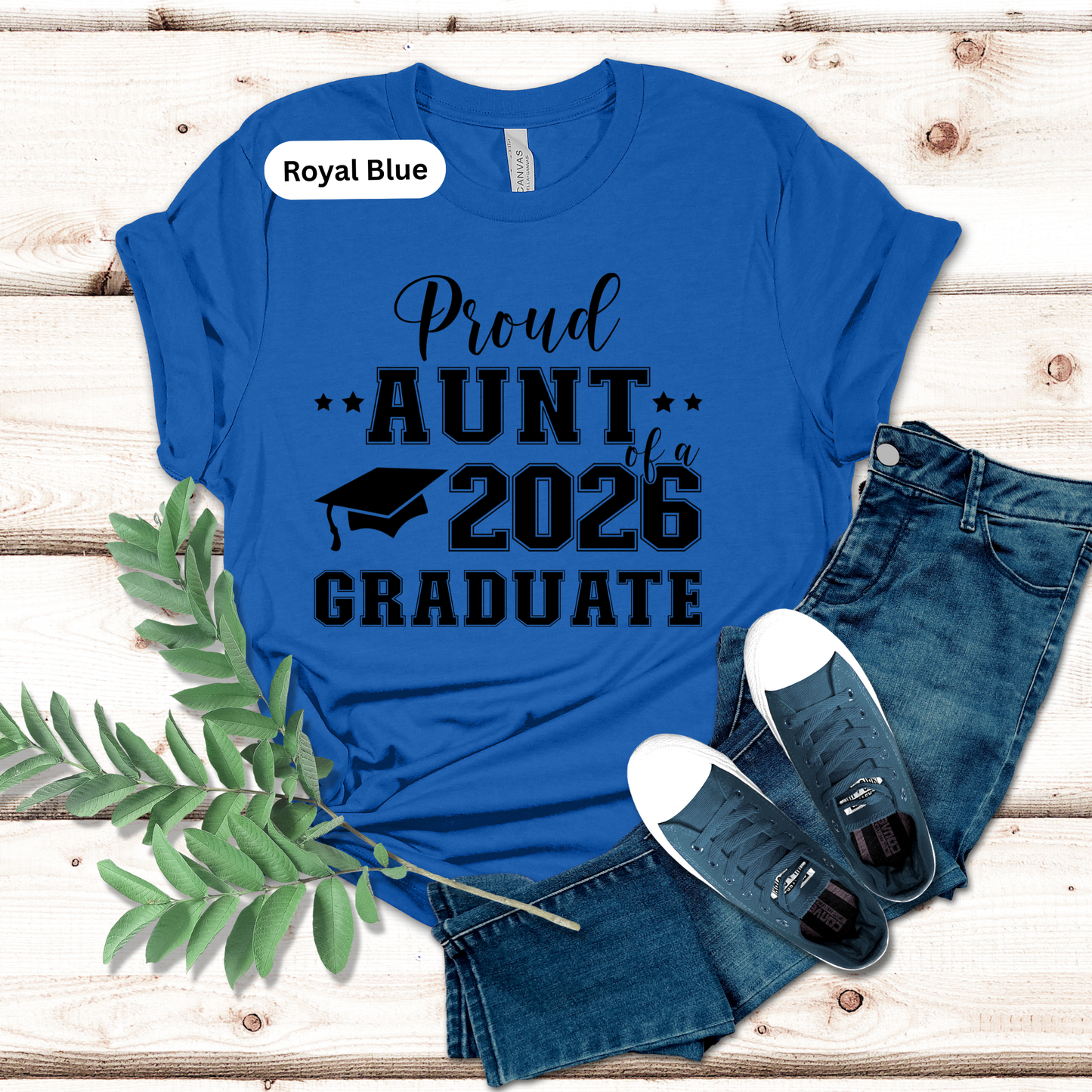 Blue t-shirt with 'Proud Aunt of a 2026 Graduate' text on a wooden surface with jeans and sneakers.