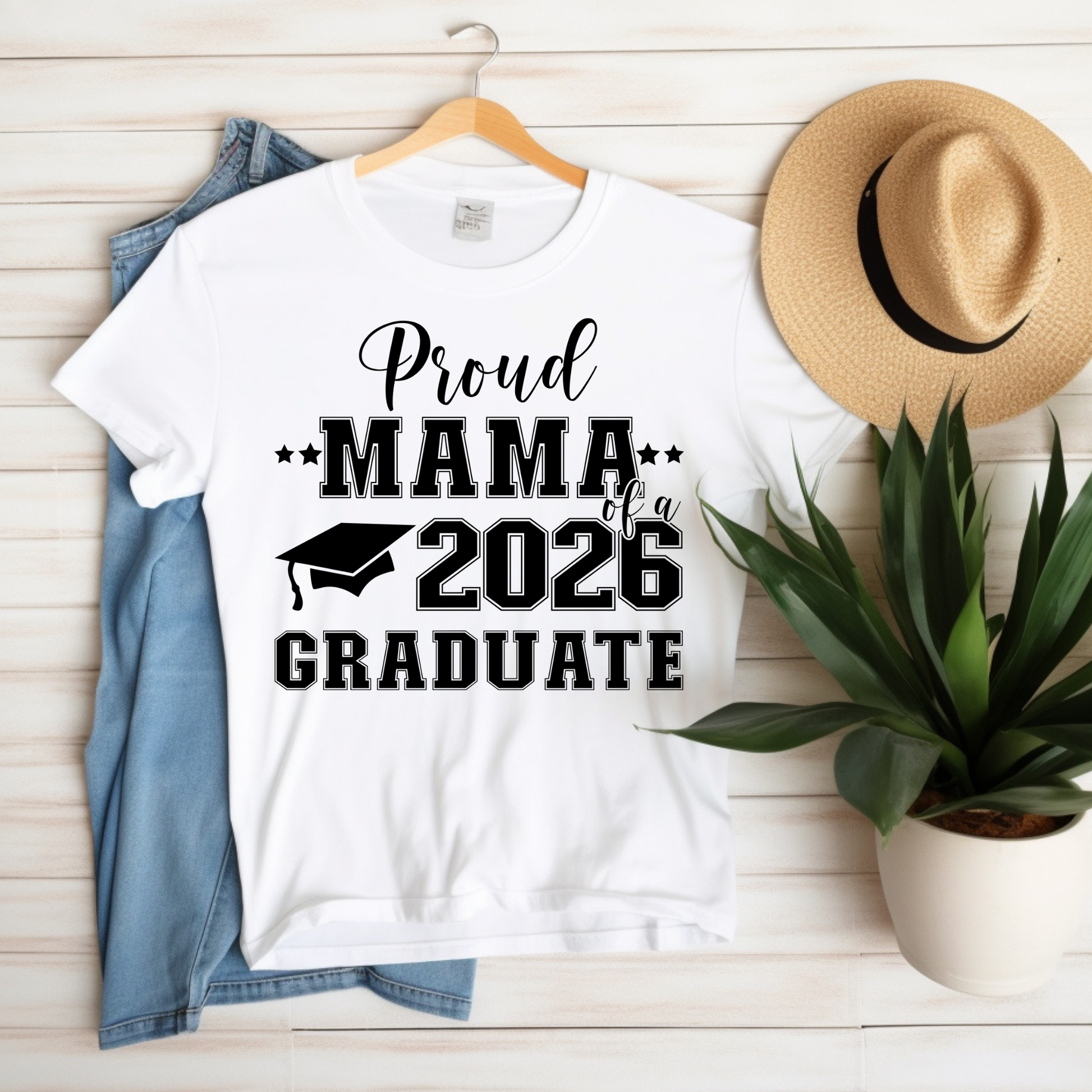 White t-shirt with 'Proud Mama of a 2026 Graduate' text on a wooden surface with a hat and plant.