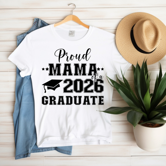 White t-shirt with 'Proud Mama of a 2026 Graduate' text on a wooden surface with a hat and plant.