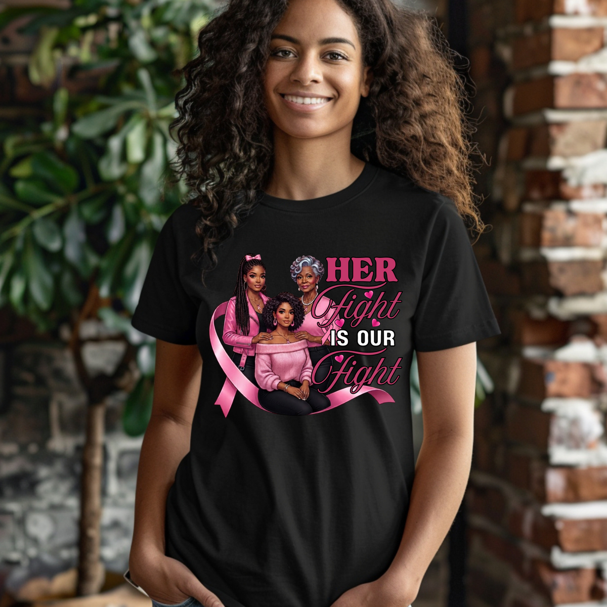 A smiling woman with curly hair wearing a black graphic t-shirt. The shirt features a pink ribbon and an illustration of three women, with the text 'HER Fight IS OUR Fight' in pink and white.