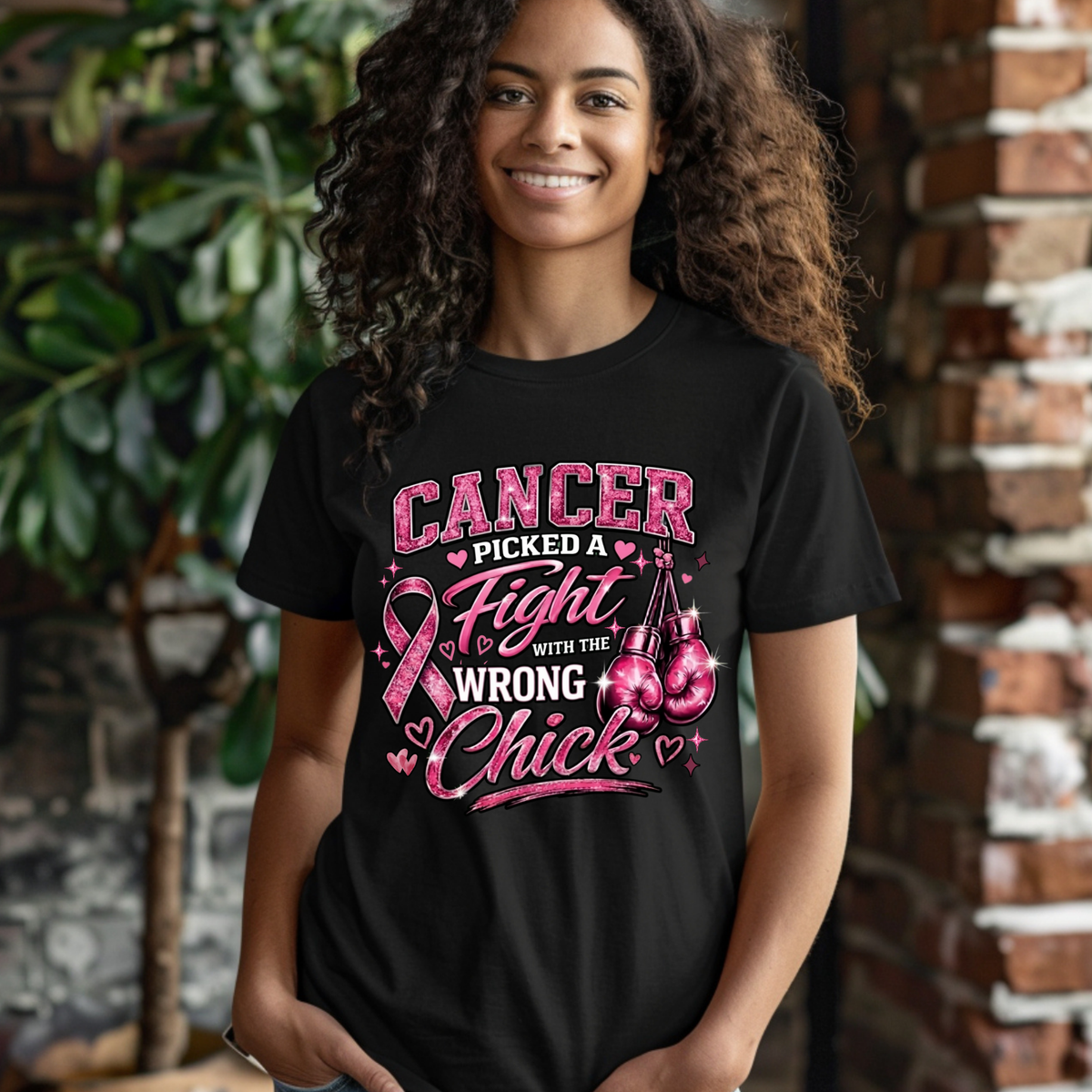 A smiling woman with curly hair wearing a black graphic t-shirt that reads 'CANCER PICKED A Fight WITH THE WRONG Chick' in pink and white, with a pink ribbon and boxing gloves illustration. She is standing in front of a brick wall and green plants.